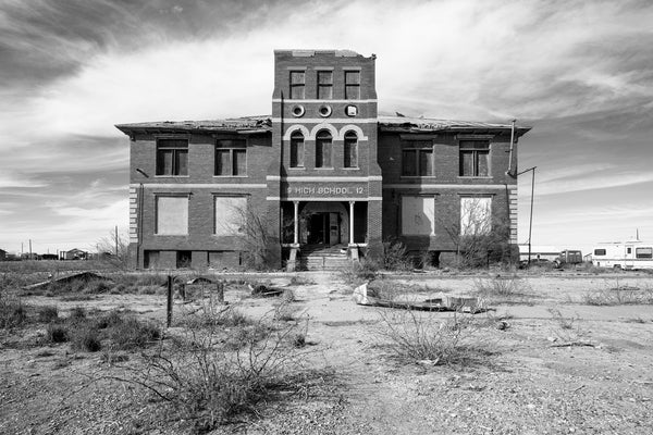 Black and white photograph of an abandoned and derelict high school building in a small town in the desert of the Western U.S. The sign over the entrance is dated 1912.
