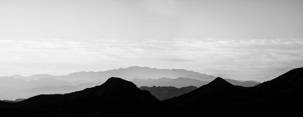 Black and white panoramic landscape photograph featuring the rising sun filtering through hazy layers of rugged western mountains.