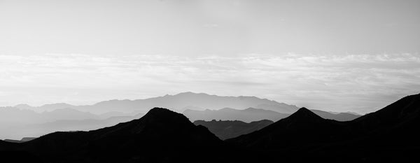 Black and white panoramic landscape photograph featuring the rising sun filtering through hazy layers of rugged western mountains.