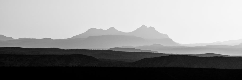 Black and white panoramic landscape photograph of layers upon layers of rugged western mountains at sunrise.