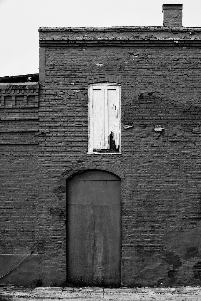 Black and white photograph of an old brick building in a small town painted all across with dark red paint, except for the white wooden shutters on a single window