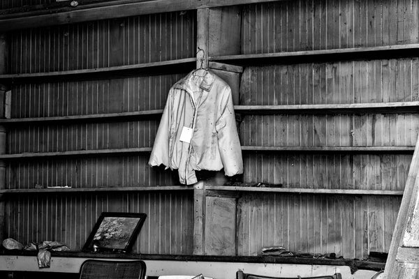 Black and white photograph of a blue jacket, with a sale tag still attached, hanging on a nail inside an old, abandoned country store.