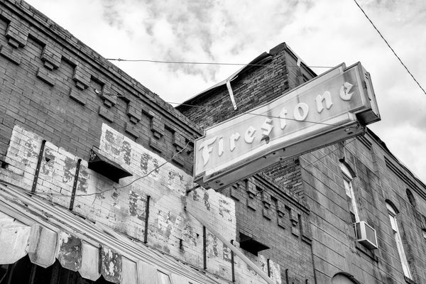 Black and white photograph of an abandoned small town hardware store with a fading painted sign that says Moore Hardware Co., and a broken plastic Firestone sign.