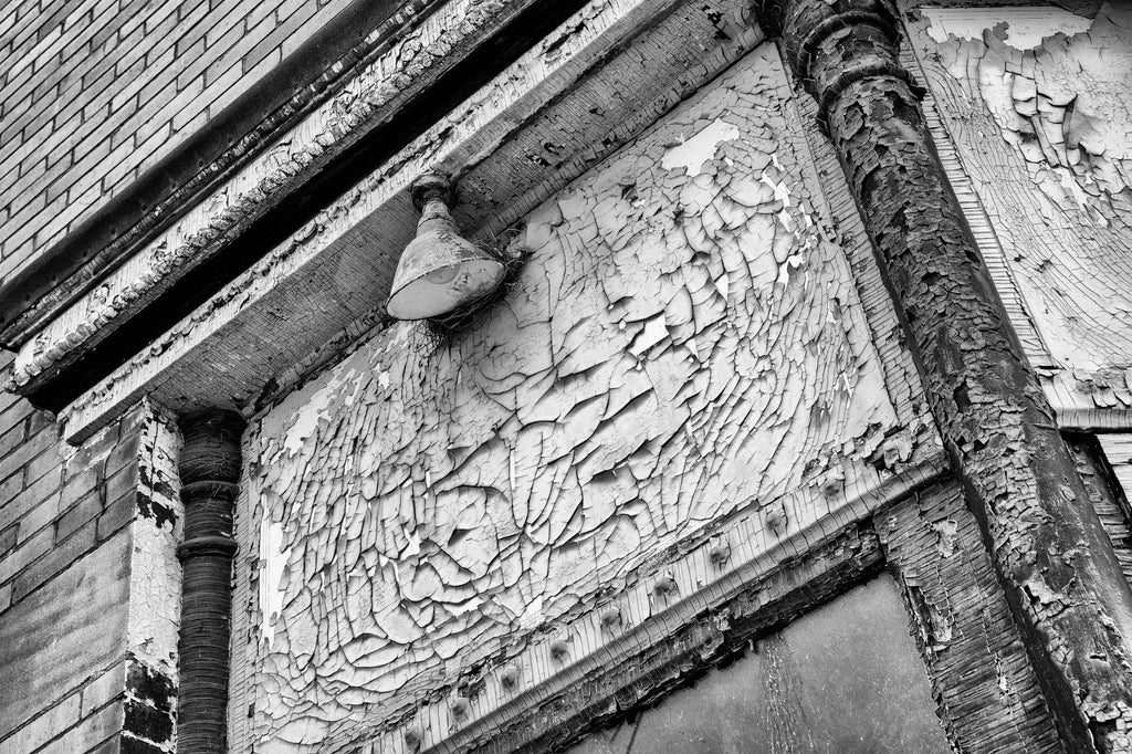 Black and white photograph of an abandoned building with a huge variety of patterns and textures in the cracked and peeling paint of its exterior. There's also a bird nest lodged behind the vintage lamp.