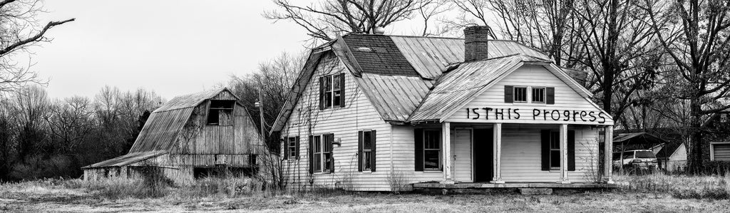 Black and white panoramic photograph showing an abandoned farmhouse with a barn in the background. The house has been marked across its front with black spray paint asking the question "Is this progress?"