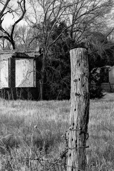 Black and white photograph of a wooden fence post near the ruins of a long-abandoned school standing among a grove of trees in rural Oklahoma.