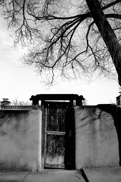 Black and white photograph of a rustic wooden door set in the gate of an adobe wall along the famous Canyon Road in Santa Fe, New Mexico.