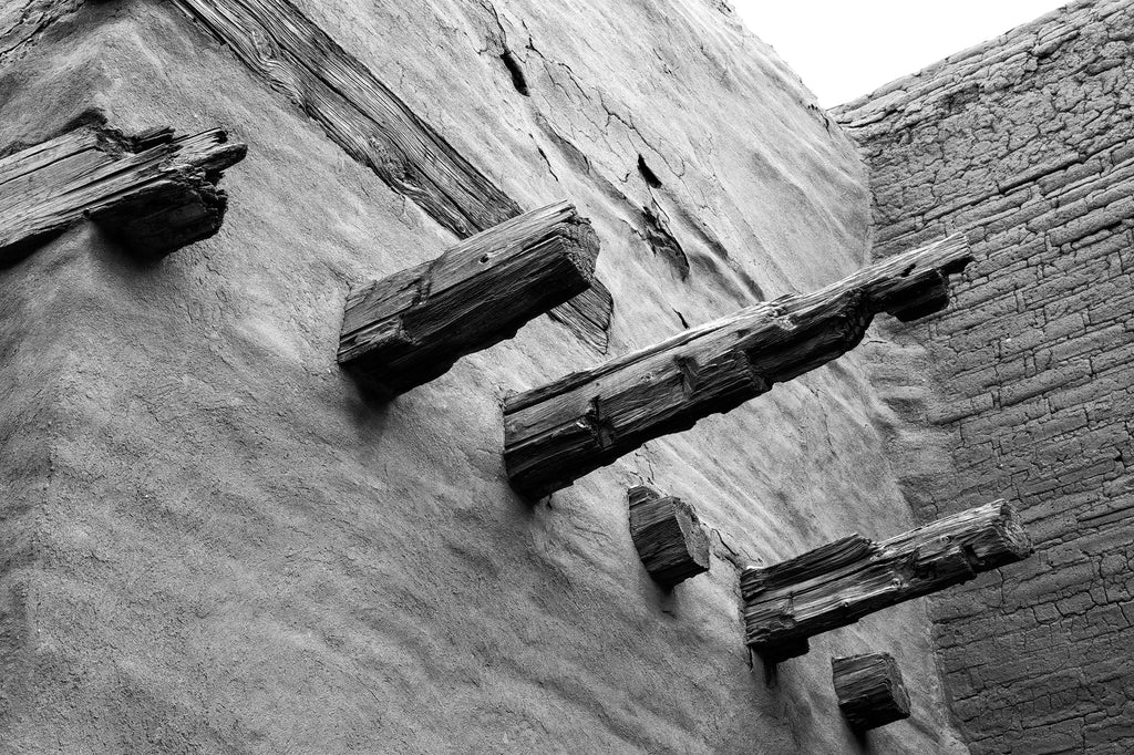 Black and white photograph inside an abandoned adobe church with weathered wooden beams and cracked bricks.