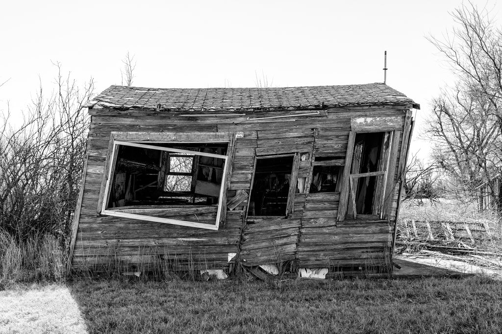 Black and white photograph of an old, abandoned wooden house that has leaned to one side, found in Texola, Oklahoma.
