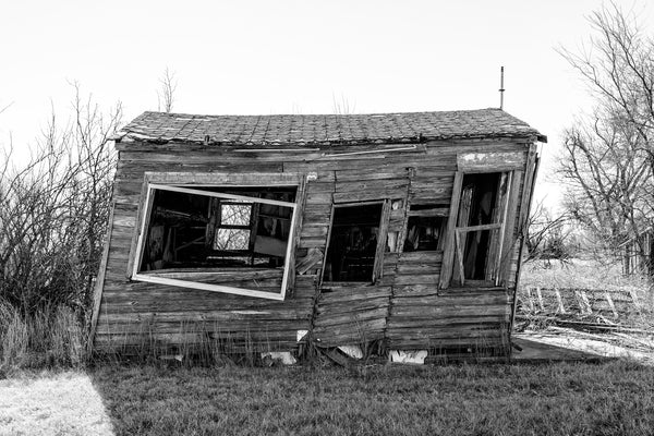 Black and white photograph of an old, abandoned wooden house that has leaned to one side, found in Texola, Oklahoma.