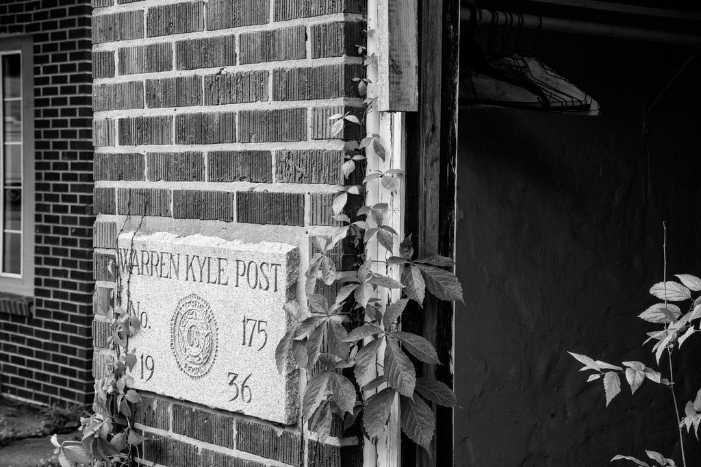 Black and white photograph of an abandoned American Legion post in a small town with its door standing open to reveal unused coat hangers inside.
