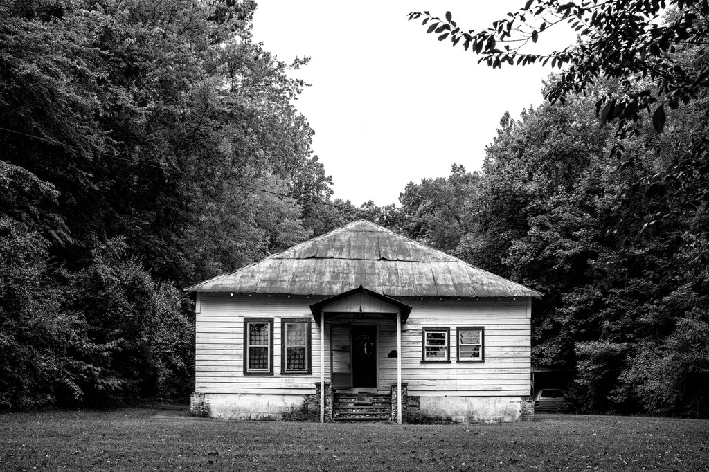 Black and white photograph of an abandoned farmhouse with a rusty metal roof and vintage automobiles parked in a homemade carport in the back.