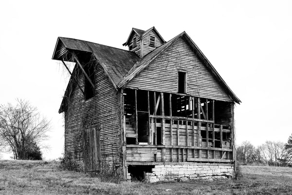 Black and white photograph of a historic abandoned old barn that once housed mules on the ground level and hay bales above. Sources say a blacksmith shop occupied an extension that's now missing leaving one side open.