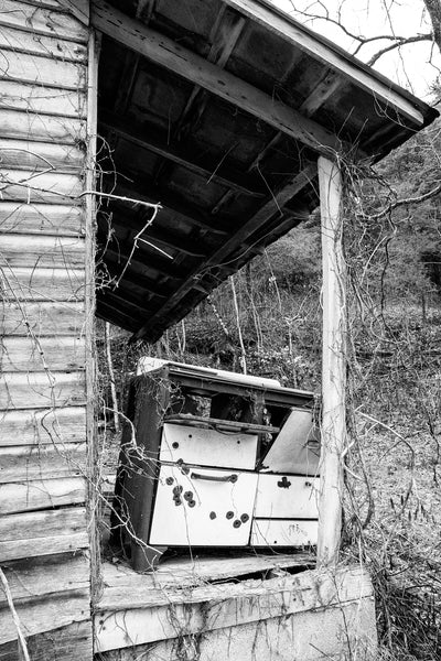 Black and white photograph of an old stove shot through with bullet holes sitting on the back porch of an abandoned farmhouse in the American South.