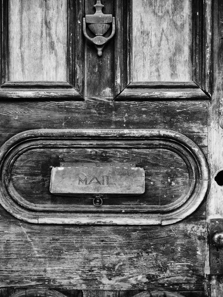 Black and white photograph of a textured, old, wooden door with a mail slot, seen in the French Quarter of New Orleans, Louisiana.