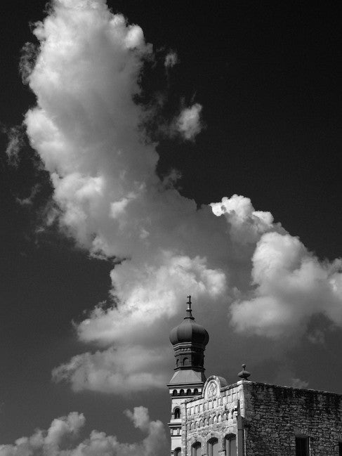 Black and white architectural photograph of a historic limestone building with a distinctive onion dome, sitting on a corner in the downtown courthouse square of Georgetown, Texas. Behind and above the architecture is a huge storm cloud, exploding upward into the deep blue sky, which has been darkened here with use of a red filter.