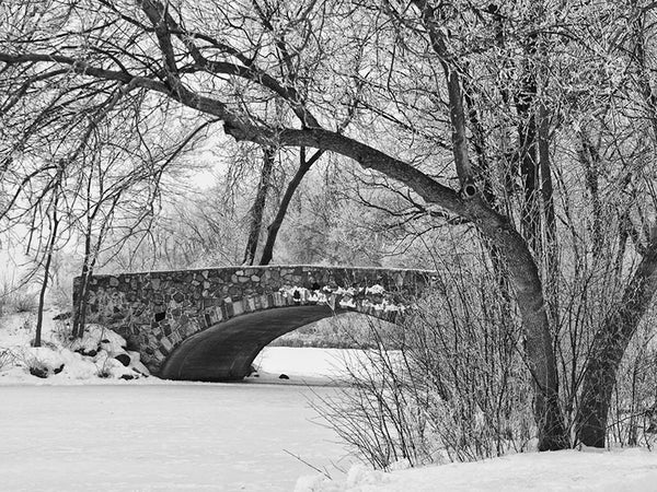 Black and white winter landscape photograph of a beautiful old stone bridge built in the 1920s, spanning a frozen pond in Madison, Wisconsin. 