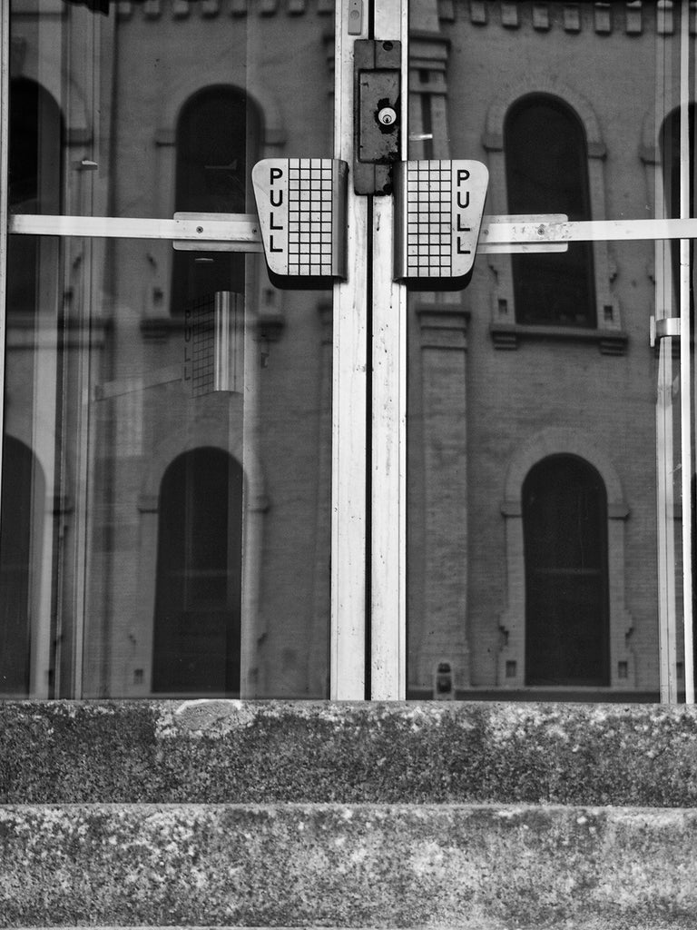 Black and white architectural detail photograph of two glass doors with retro "pull" door handles, seen in urban Milwaukee.