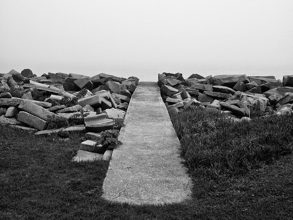 Black and white landscape photograph of a sidewalk leading to nowhere, but it actually goes to the shore of Lake Michigan, which is hidden in a thick blanket of early morning fog.