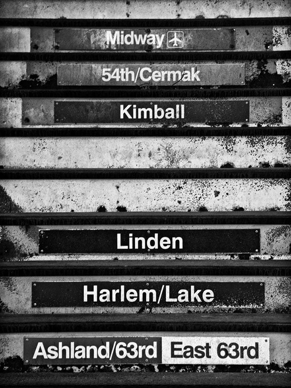 Black and white photograph of the a well-worn metal staircase to the elevated train platform in Chicago. The steps include destination names.