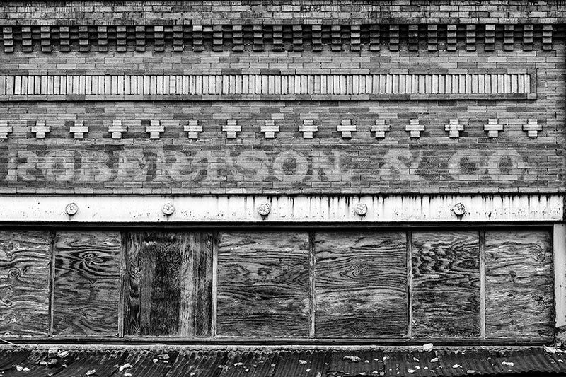 Black and white photograph of the abandoned Robertson & Co. building in the deserted former downtown of Adams, Tennessee. This photograph shows the name of the business in fading white paint on the bricks of the storefront.