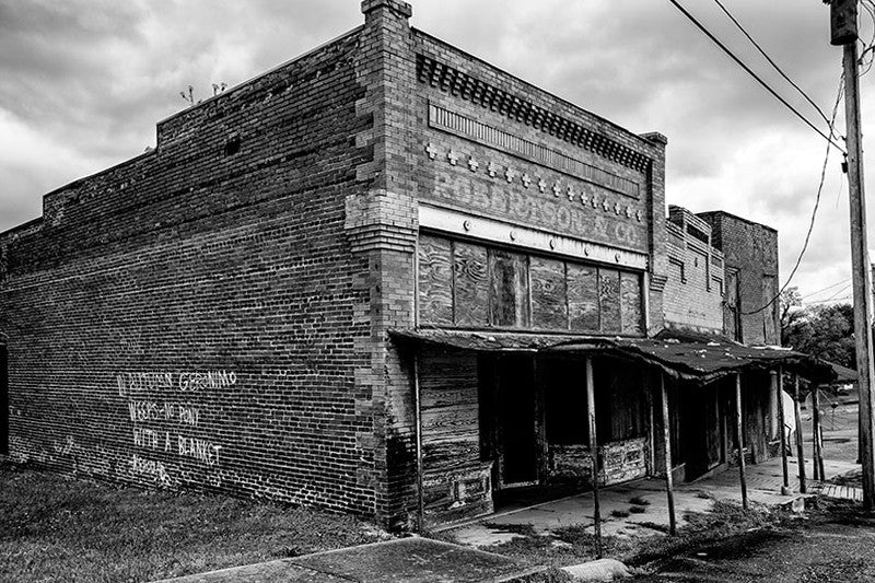 Black and white photograph of the abandoned Robertson & Co. building in the deserted former downtown of Adams, Tennessee. On the side wall of the building, someone spray-painted a quote from Jack Kerouac, "In Autumn Geronimo Weeps — No Pony with a Blanket."