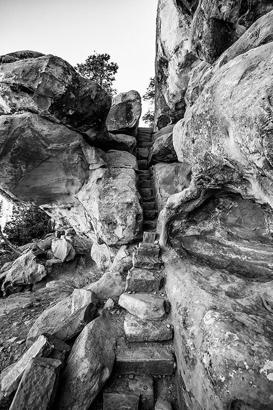 Black and white fine art photograph of one of the rugged hiking trails at Mesa Verde in Colorado. The path travels under huge rock ledges, between massive boilders and walls, past the ancient petroglyphs, and up a climb worthy of a mountain goat.