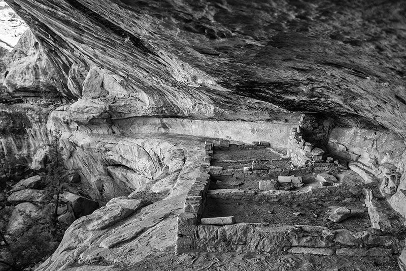 Black and white photograph of the foundations of a small Pueblo cliff dwelling on a low ledge at Mesa Verde, Colorado.