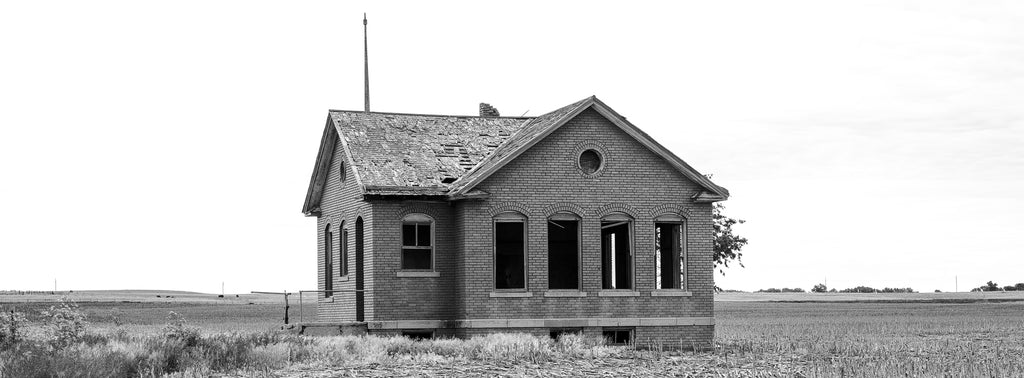 Black and white wide-format photograph of a red brick one-room schoolhouse built in 1920 along an unpaved country road on the Kansas prairie.