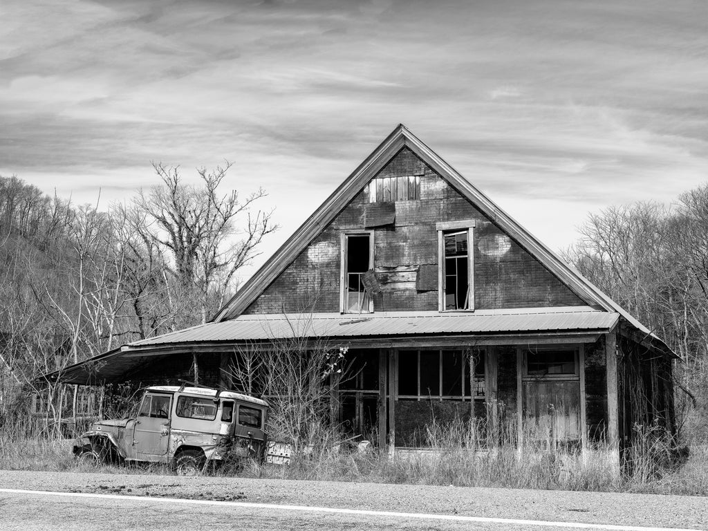 Black and white photograph of an abandoned wooden general store in the American South with a broken down vehicle parked in the tall grass by the road.
