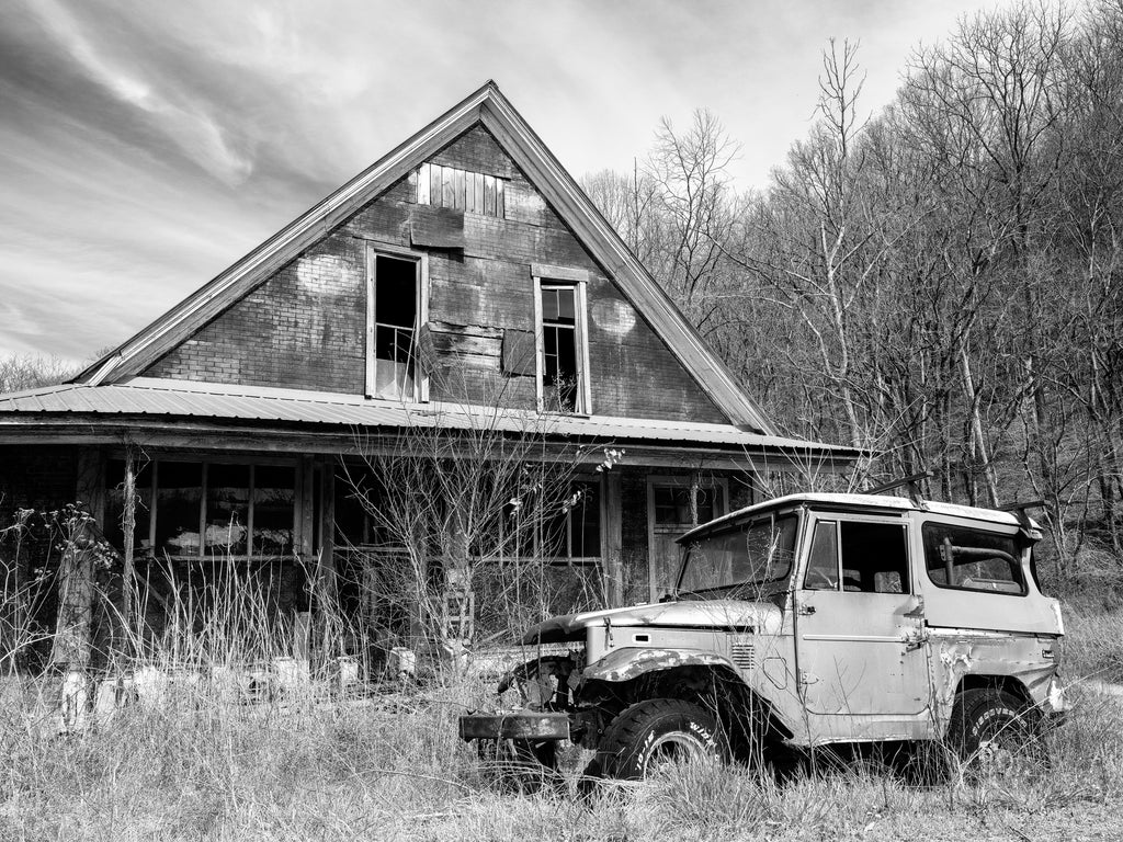 Black and white photograph of an abandoned wooden general store in the American South that has a broken-down early-1980s model Toyota Land Cruiser deserted in the tall grass by the road.