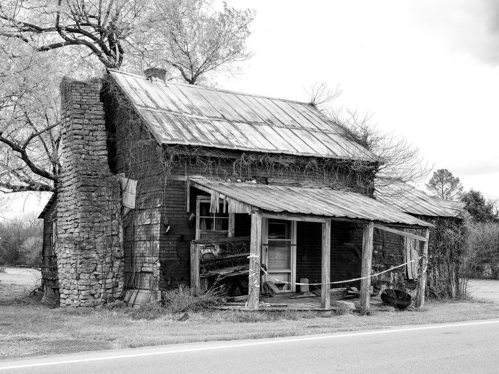 Black and white photograph of an abandoned house in the rural American South that has a weathered old piano sitting on the front porch.