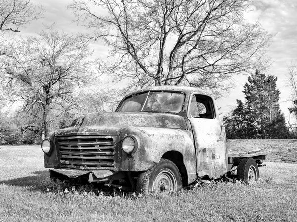 A black and white photograph of a weathered, rusted vintage flatbed truck parked in a grassy field with bare trees in the background.