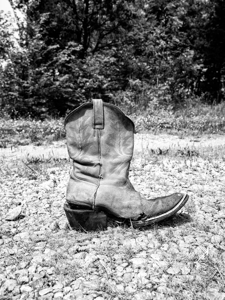 Black and white photograph of an Old cowboy boot on a gravel path with trees in the background