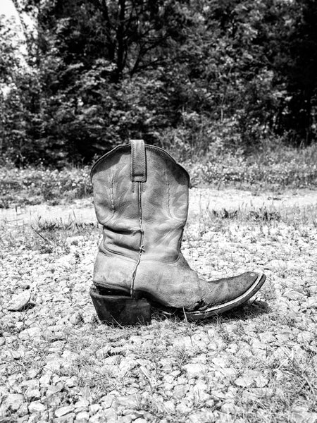 Black and white photograph of an Old cowboy boot on a gravel path with trees in the background