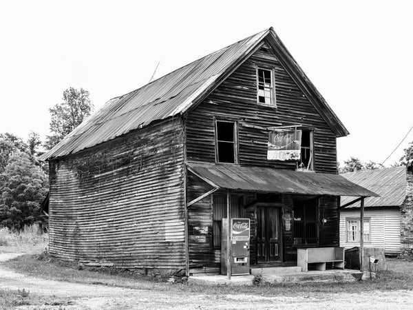 Black and white photograph of an abandoned wooden general store called Woody's, located in Auraria, Georgia. The old store still features a rusty Coca-Cola sign hanging over the entrance, and a vintage soft drink machine on the porch.