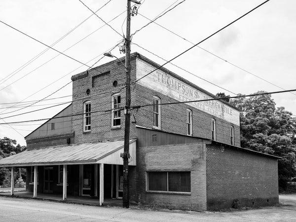 Black and white photograph of a vacant old mercantile store in the small town of Haralson, Georgia, known for being a filming location in the TV show The Walking Dead. A large sign on the brick wall says "Thompson's Mercantile, Feed Seed Supply."