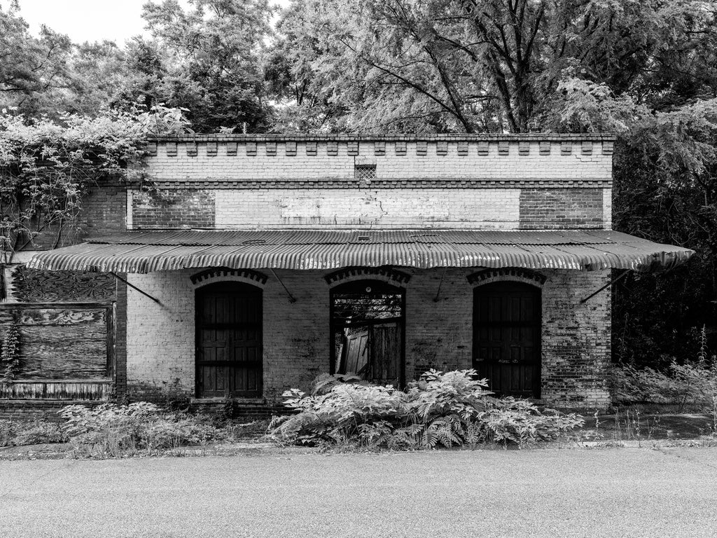 Black and white photograph of an abandoned historic storefront that once served as Starke's Feed Store, a farm supply store in the small town of Seale, Alabama. It sits on a street where every building is now vacant and in ruins.