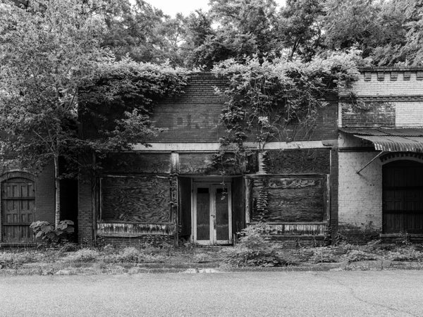 Black and white photograph of an abandoned historic storefront that once served as City Drug Store with a pharmacy and soda fountain, in a small community Seale, Alabama. It sits on a street where every building is now vacant and in ruins.