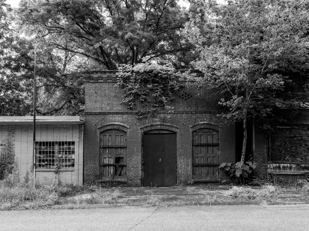 Black and white photograph of an abandoned historic storefront that may have served as a hardware store in a small community in the American South. It sits on a street where every building is now vacant and in ruins.