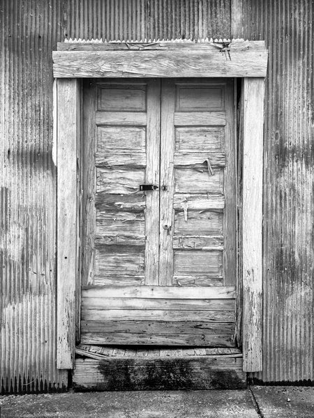 Black and white photograph of textured and weathered wooden doors on the corrugated steel wall of a big cotton warehouse in the American South.