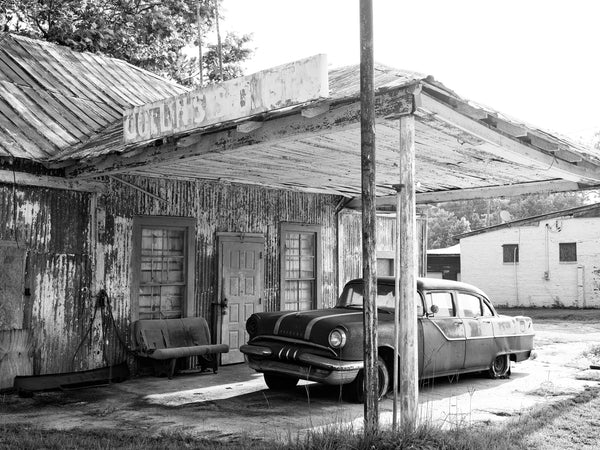 Black and white photograph of a 1950s-era Pontiac parked under the awning of an abandoned old service station in the American south.