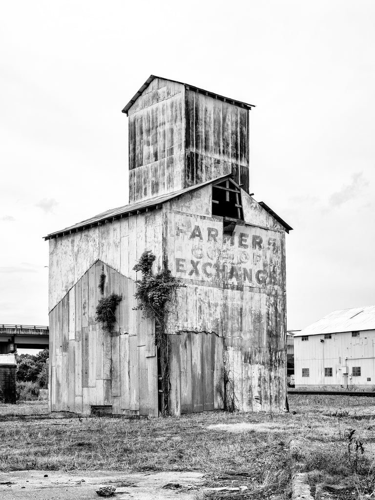 Black and white medium format photograph of an abandoned grain elevator with a faded sign saying Farmers Co-Op Exchange on its side in the small town of Paragould, Arkansas.