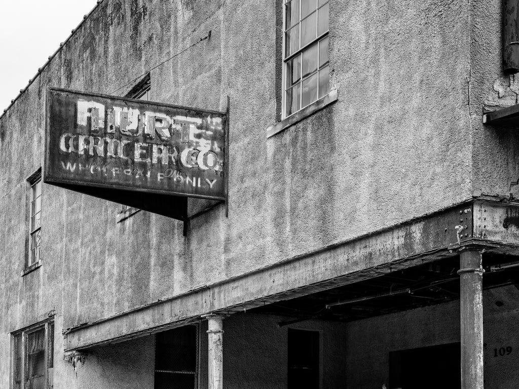 Black and white photograph of a beautifully layered old sign on the exterior of the Hurt Grocer Company, a wholesale grocery service that once operated in Paragould, Arkansas. Online sources suggest it was built in 1920.