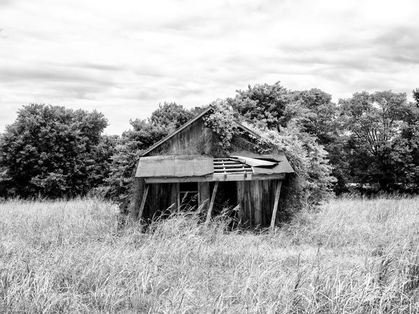 Black and white photograph of an old wooden house with leaning porch posts standing abandoned in a grassy field in the American south.