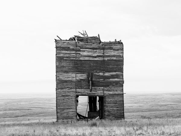 Black and white photograph of a wooden grain elevator with its top section missing as seen in the small community of Okaton on the high plains of South Dakota.