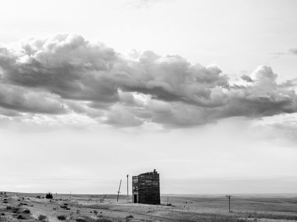 Black and white photograph of a broken wooden grain elevator standing under a vast storm cloud in the small community of Okaton on the high plains of South Dakota.