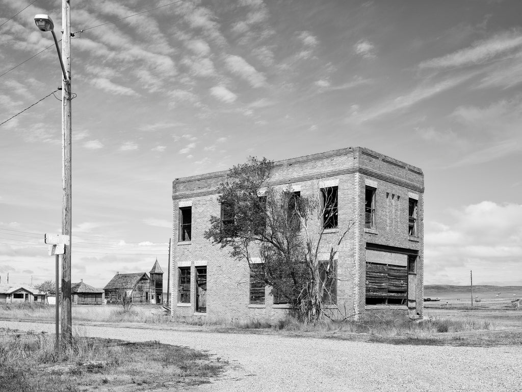 Black and white photograph of an abandoned building on a street corner in the nearly-a-ghost-town at Cottonwood, South Dakota. More abandoned buildings including an old wooden church can be seen in the background.