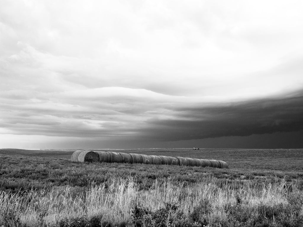 Black and white landscape photograph of the dark clouds of a storm front moving over farmland in the vast landscape near the Badlands of South Dakota.