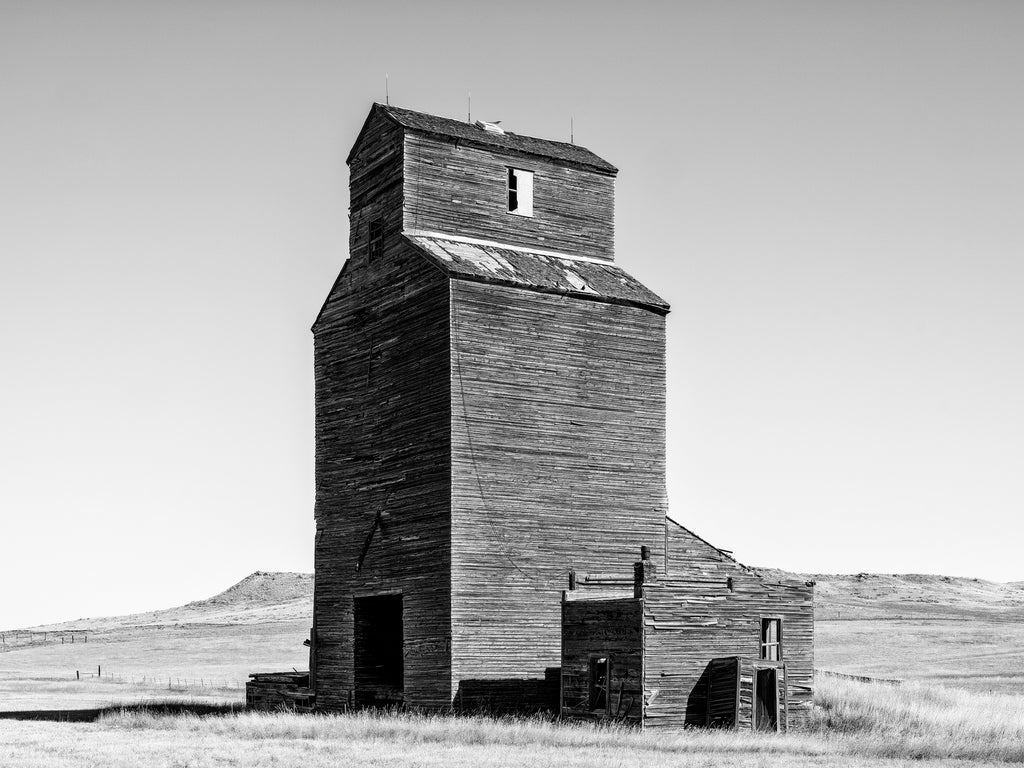 Black and white photograph of an abandoned wooden grain elevator standing on the rolling prairie of Montana.